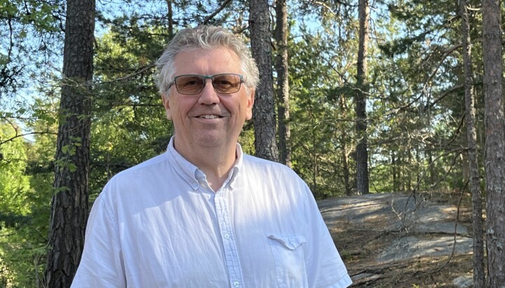 På bilde: NRK-veteran Geirmund Henjum står i skogen og smiler med solbriller på.