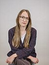 Kvinne med briller og langt hår sitter foran en lys bakgrunn.