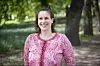 Smilende kvinne i rosa mønstrete topp står i en park med trær og grønt bakteppe.