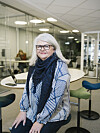 Kvinne med briller og blått skjerf sitter i et lyst kontor med glassvegger og bord.