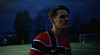 Fotballspiller i rød og svart drakt står på en mørk fotballbane ved et mål.
