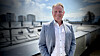 Smilende mann i lys blazer står på takterrasse med by og vann i bakgrunnen.