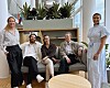 Fem personer står og sitter samlet i et lyst rom med sofa, planter og store vinduer.