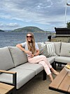 En kvinne sitter i en lys sofa på en terrasse med sjøutsikt og fjell i bakgrunnen.