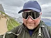 Person i caps og solbriller tar selfie på en fjellsti med fjell og dal i bakgrunnen.