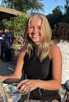 Smilende ung kvinne sitter ved et utendørs bord med glass og mobiltelefon.