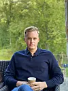En mann sitter i en stol ute og holder en kaffekopp foran seg, med grønne trær i bakgrunnen.