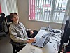 Person sitter ved et skrivebord med datamaskin, tastatur og kabler i et lyst kontor.